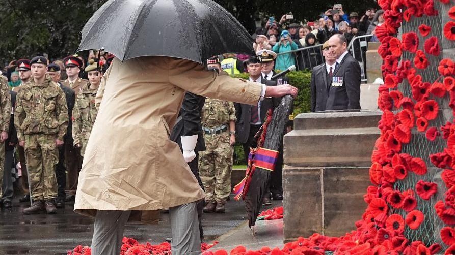 King Charles wearing a brown coat and holding a black umbrella lays at wreath a war memorial decorated with red poppies. Soldiers and the public line the background of the photo.