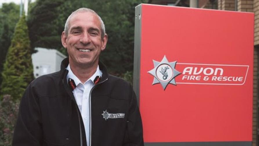 Simon Shilton is pictured smiling at the camera, wearing an Avon Fire and Rescue fleece, next to a large sign for the organisation. He has short dark hair, and is standing outside what appears to be the service's headquarters. 