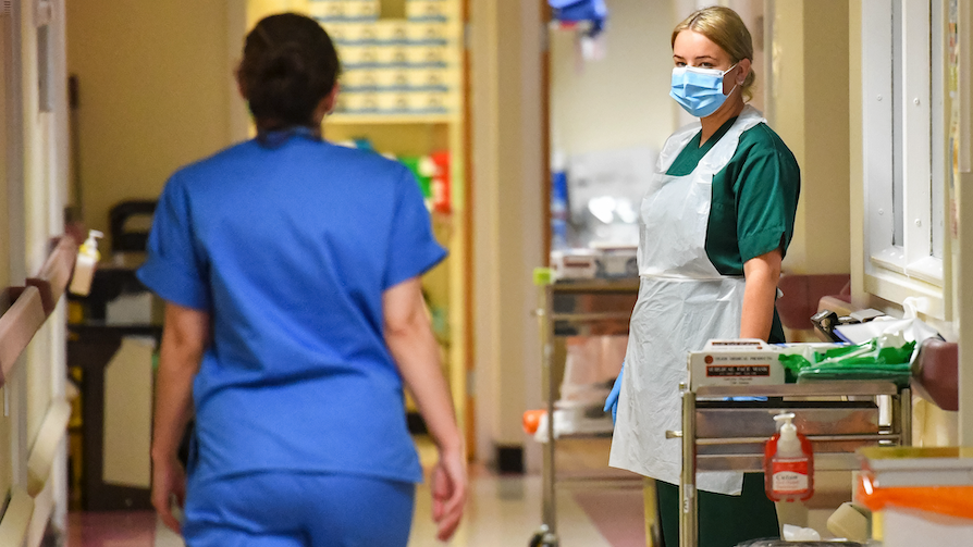 Two women in a hallway in a medical setting wearing face masks and blue scrubs.