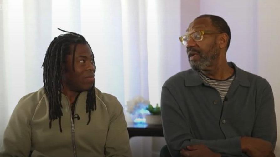 Ade Adepitan, wearing a light green fleece top, is sat next to Sir Lenny Henry, who is wearing glasses and a grey shirt over a striped t-shirt.