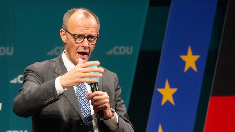 Friedrich Merz gives a speech - wearing a grey suit against a light blue background