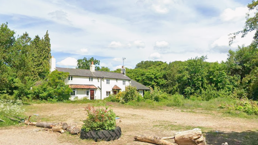 A two-storey building, largely painted white, with a large patch of open land in front of it and large trees and a big tyre in front of it to prevent it being used as a car park.