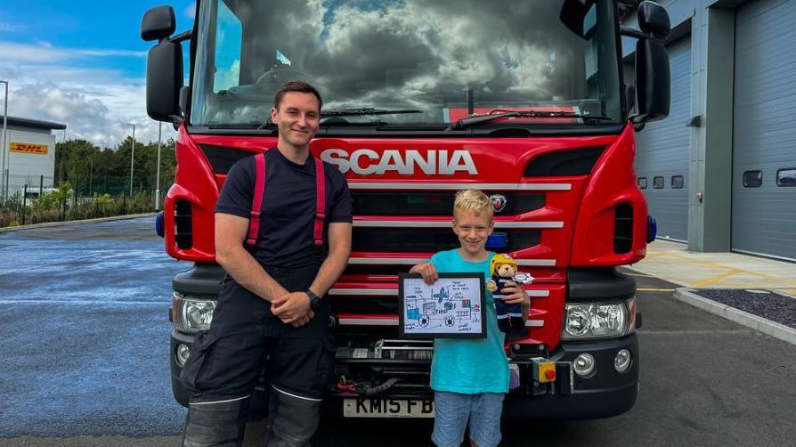 A firefighter in a blue T-shirt, with red braces and blue trousers tucked into boots. He is standing beside Luca, a primary-school-aged boy, who is holding up his hand-drawn fire engine design. Both are standing in front of a bright red fire engine and smiling.