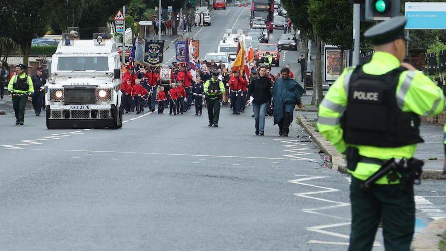 Police in the forground with their back to the camera. In the background a parade of people in red uniform led by a police land rover