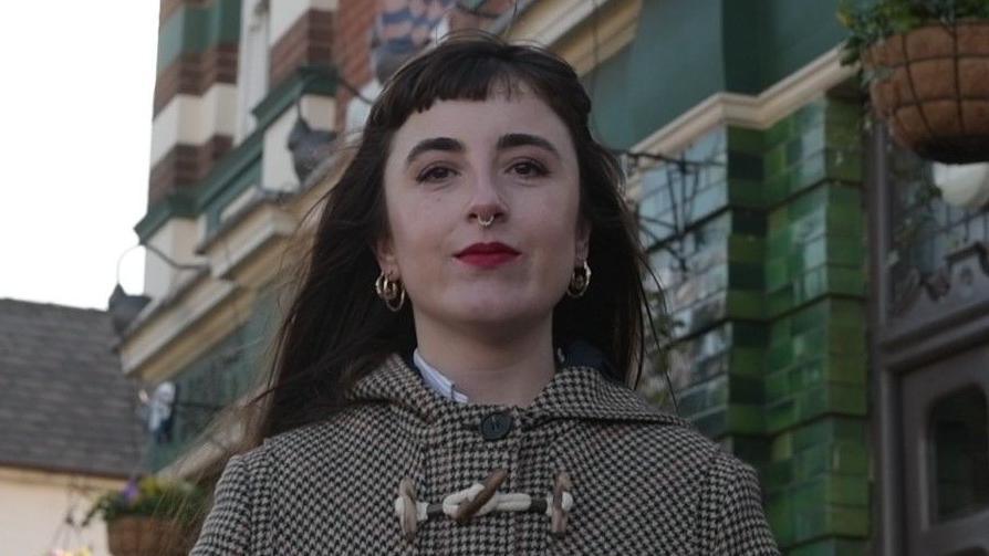 Hayley standing outside the Painters Arms in High Town. She has long straight brown/reddish hair with a short fringe, wears a nose ring and has bright red lipstick on.