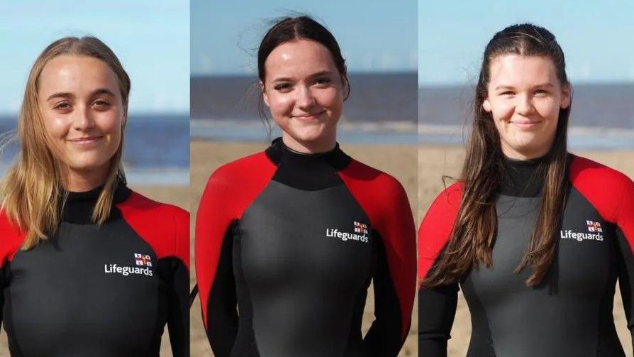 Professional photos of RNLI lifeguards Lois Kemp, Katie Roscoe and Effie Kennedy standing on the beach in black and red lifeguards outfits and smiling into the camera
