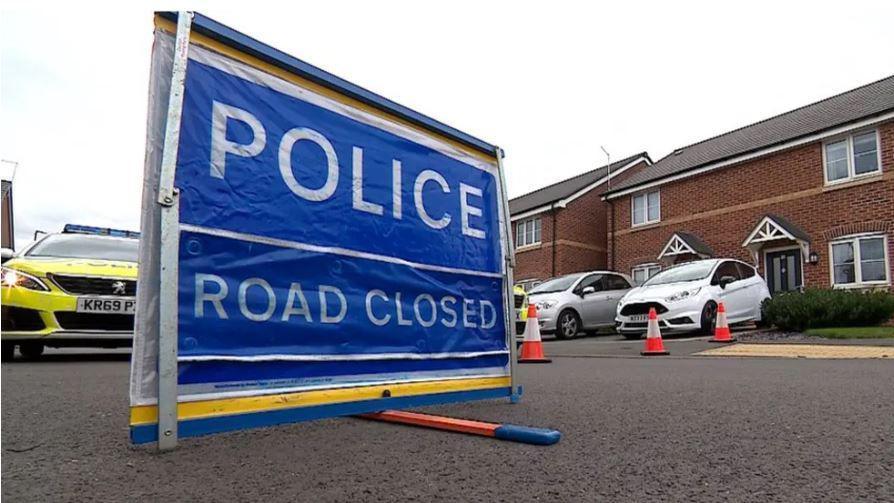 A "police road closed" sign with the back drop of houses and a police car on a street