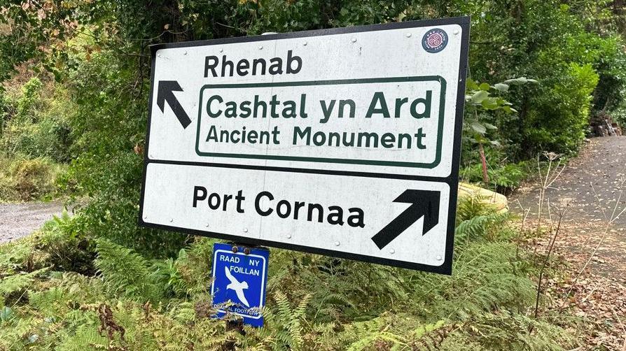 A large white sign with Port Cornaa and an arrow pointing right below another arrow indicating left for Cashtal yn Ard.