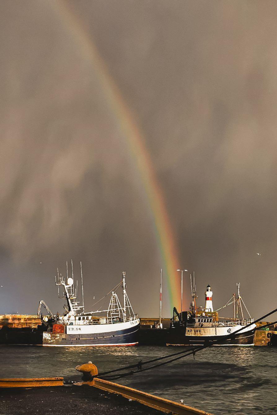 Two dark blue and white fishing trawlers in harbour, with a rainbow arc in a dark sky behind them, and a red and white lighthouse.