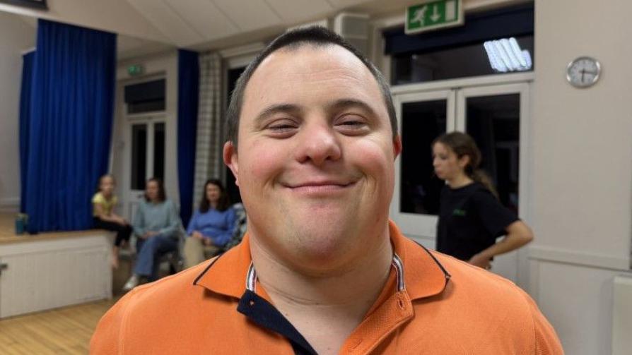 Nathan, who has Down's Syndrome, smiles for the camera. He is wearing an orange polo shirt and has very short black hair. Other class members and parents can be seen in the background in the village hall.