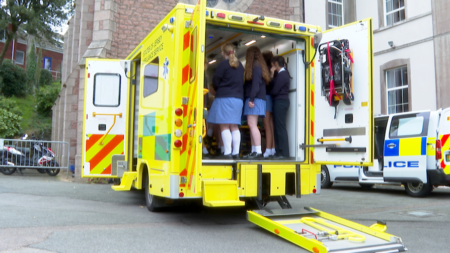 A yellow ambulance with students in it looking around, parked on a Tarmac car park with buildings in front, a police van on the right beside it