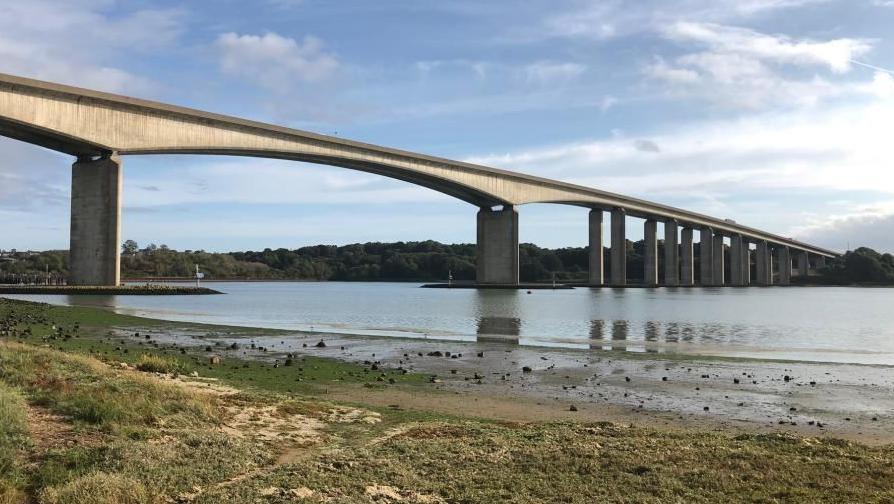 The Orwell Bridge, over the River Orwell near Ipswich: a grey stone bridge spanning a grey river. A grass and mud ban is in the foreground. Parts of the bridge are also reflected in the water,