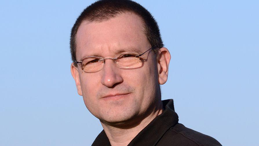 David Johnson looking at the camera and wearing a black shirt and glasses. Behind him is a light blue background.