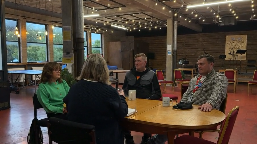 Two women and two men, including Danny Woodburn, sit round an oval wooden table in a large open plan office. The area is sparsely furnished, but some red dining style chairs and a couple of tables can be seen. There is a stage at the back with music stands on it. One woman in a dark jumper has her back to the camera. To her left is a woman with glasses wearing a green buttoned-up cardigan. On the other side of the table is a man with light brown hair wearing a black coat, and to his left Danny sits with one hand on the table.