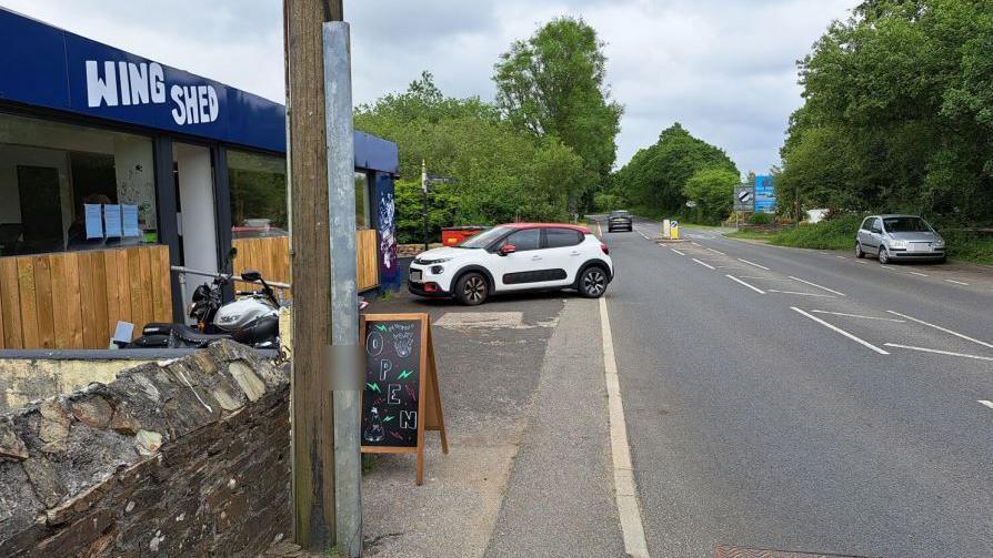 A white car with a red roof parked outside Wing Shed in London Apprentice. It is blocking the pavement and has part of its rear end edging into the road. A blackboard sign saying 'open' is on the roadside.