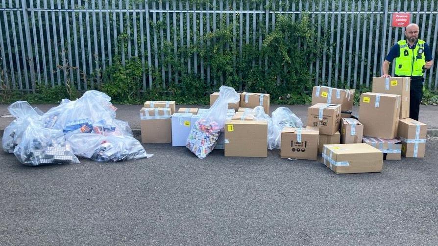 A police officer, standing by a large number of cardboard boxes, bags of illegal vapes and cigarettes, by a metal fence, with greenery growing up it. The man is standing to the right.