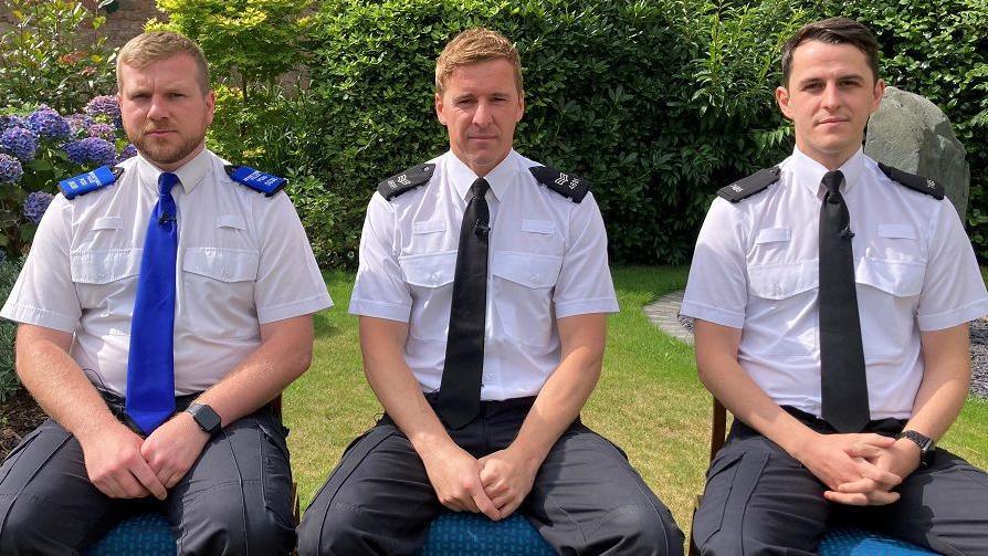 PCSO Tim Parry, Sgt Greg Gillespie and PC Luke Holden are sitting down on chairs in a garden. They are all wearing their police uniform. PCSO Parry is wearing a blue tie and epaulettes on his shoulders. Sgt Gillespie and PC Holden are wearing black ties and epaulettes on their shoulders.