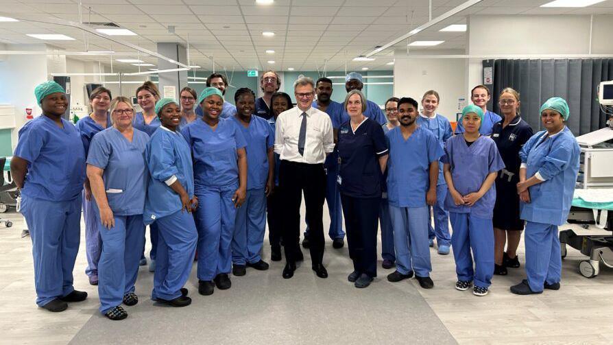 A group picture on the ward. Hospital staff are wearing blue scrubs, Mr Wood is wearing a white shirt, black trousers and a tie.