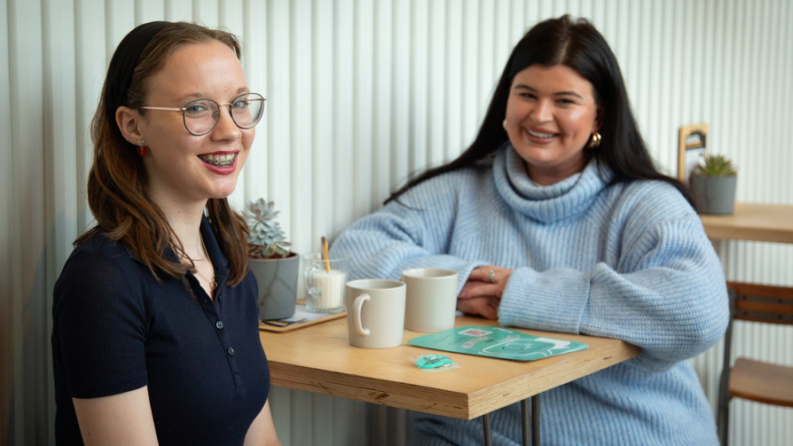 Kitty and Jessie sat at a table in a coffee shop. Kitty has long dark hair and is wearing a black headband. She has glasses and braces and is wearing a navy t-shirt. Jessie has long black hair and gold hoop earrings, and is wearing a light blue knitted roll neck jumper. 