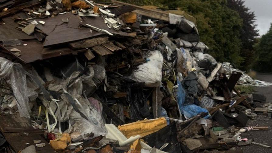Tree-lined road with vast pile of fly-tipped rubbish strewn to the left side.