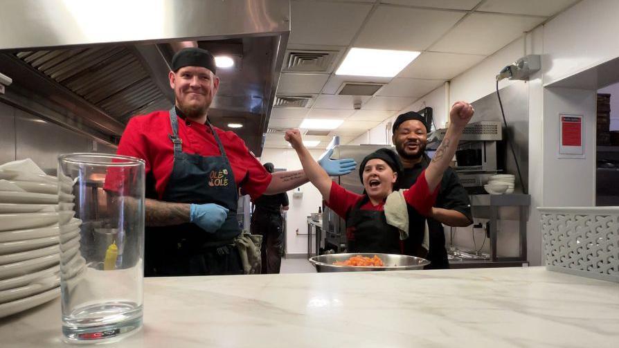 Two male kitchen staff flank a female colleague with her arms raised in celebration in the kitchen of the Olé Olé restaurant. They wear red and blue uniforms and blue chefs hats.