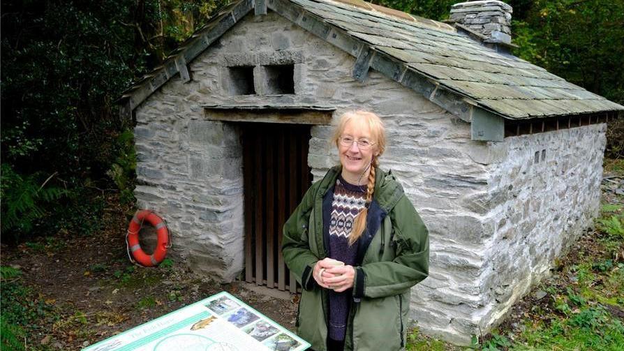 Pam Mills is standing in front of the restored fish house. It is a small rectangular stone-built building with a slate roof