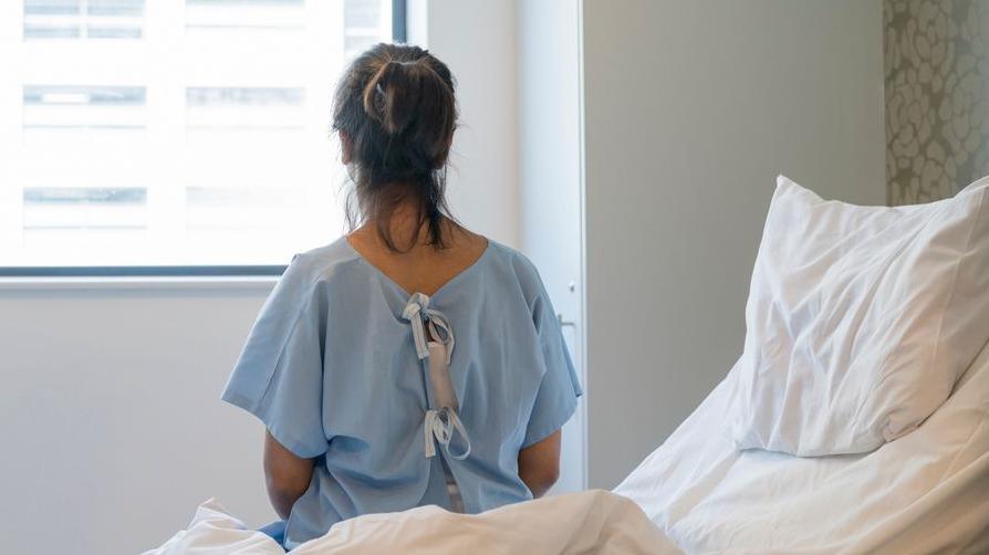 A woman wearing a hospital gown sitting on a hospital bed looking out of a window. 