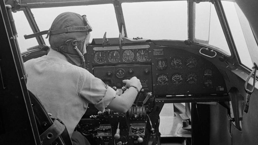 A World War Two pilot wearing head gear and goggles at the controls of an aircraft. 