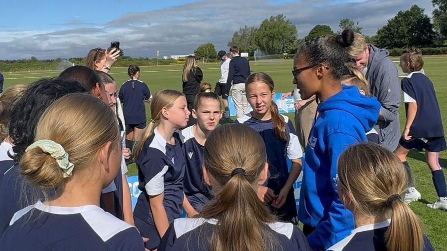 Rachel Yankey speaking to a group of girls during her visit to an Oxford football training session run by Oxford United for Sport England backed campaign Play Their Way. It is cloudy.