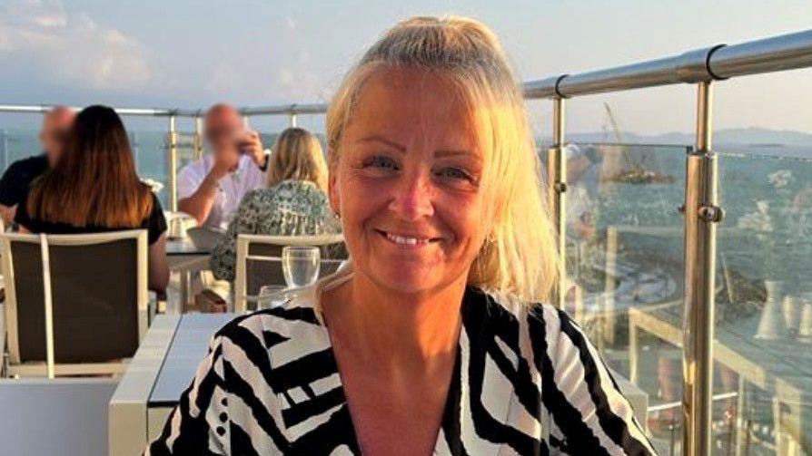 Anita Rose sitting at a table in the open air on a veranda overlooking a urban landscape. She is wearing a black and white patterned top and is smiling.