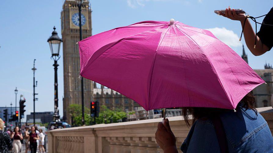 A person shielding from the Sun under an umbrella with the Houses of Parliament in the background