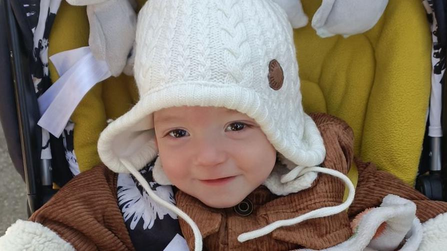 Charlie Roberts, a young boy with brown eyes. He is sitting in a pushchair with a brown corduroy jacket and white woollen hat.