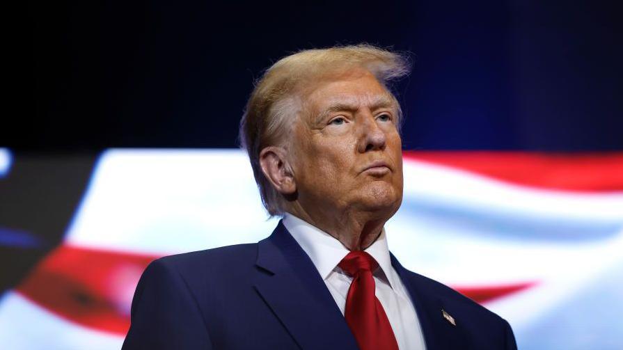 Donald Trump looks ahead with a serious expression on his face. He is wearing a navy suit, white shirt and red tie.