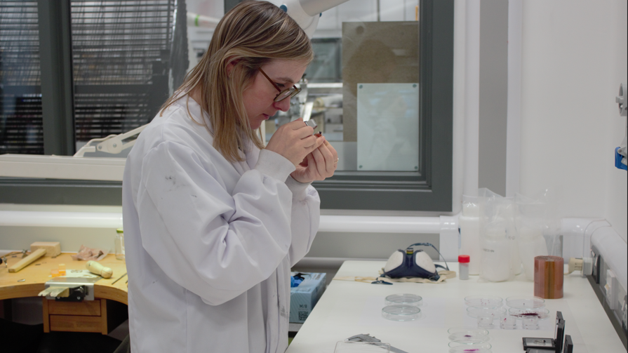 Sofie Boons studies a gemstone in her laboratory. She is wearing a white lab coat and glasses