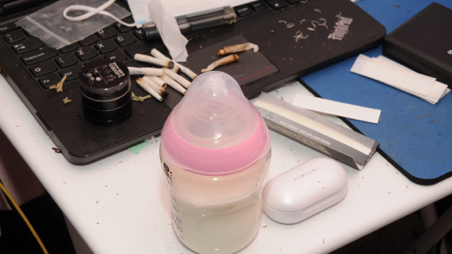 A baby's bottle filled with milk sits on a desk, next to a computer keyboard, covered in cannabis joints and cigarette papers.