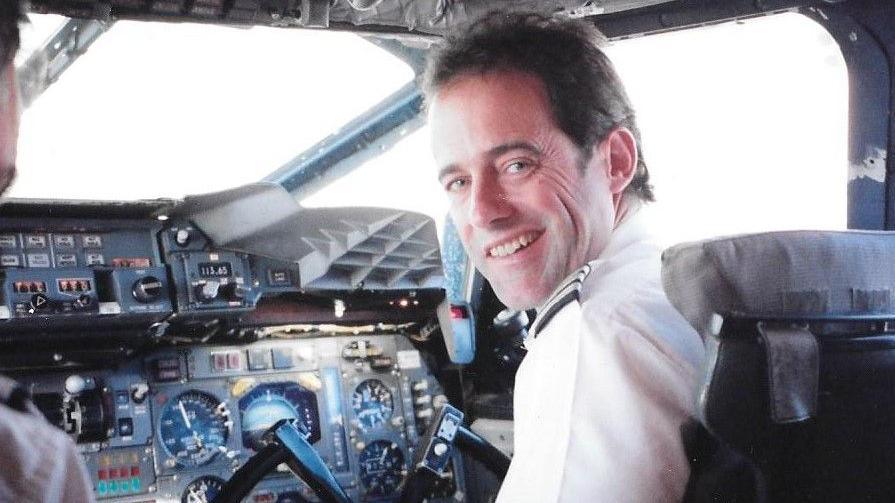 A image of John in the cockpit as a young pilot. He is sitting in the pilot's seat, with the steering wheel in front of him, dressed in a white shirt.
