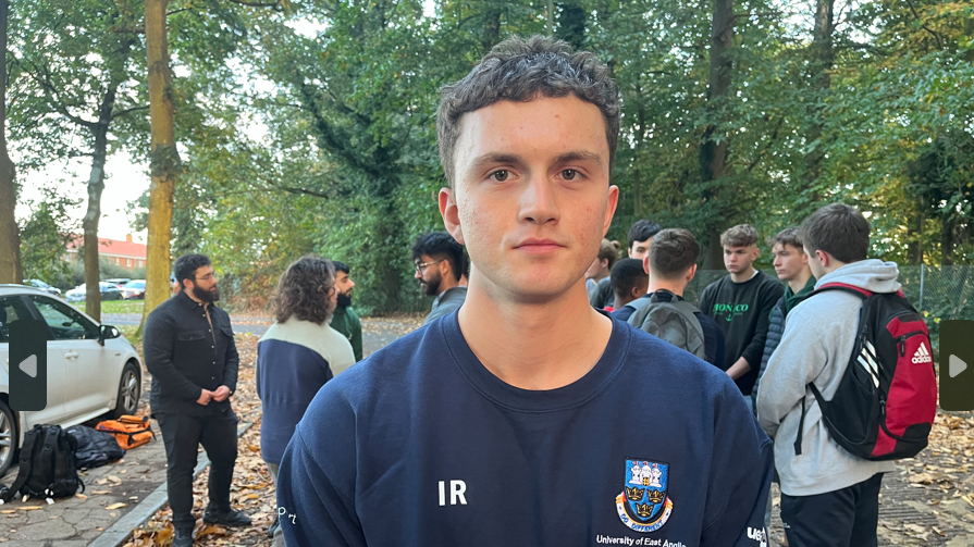 Isaac Rodriguez is looking directly at the camera with a solemn expression. Behind him is a group of students conversing. Mr Rodriguez is wearing a navy University of East Anglia branded sweatshirt. It has the university logo on the top right and the letters IR on the top left. Mr Rodriguez has brown eyes and short brown hair with a slight curl in it. 