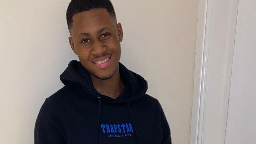 A man with short black hair and a dark-coloured hooded jumper smiles as he leans slightly to his right in front of a cream-coloured wall.