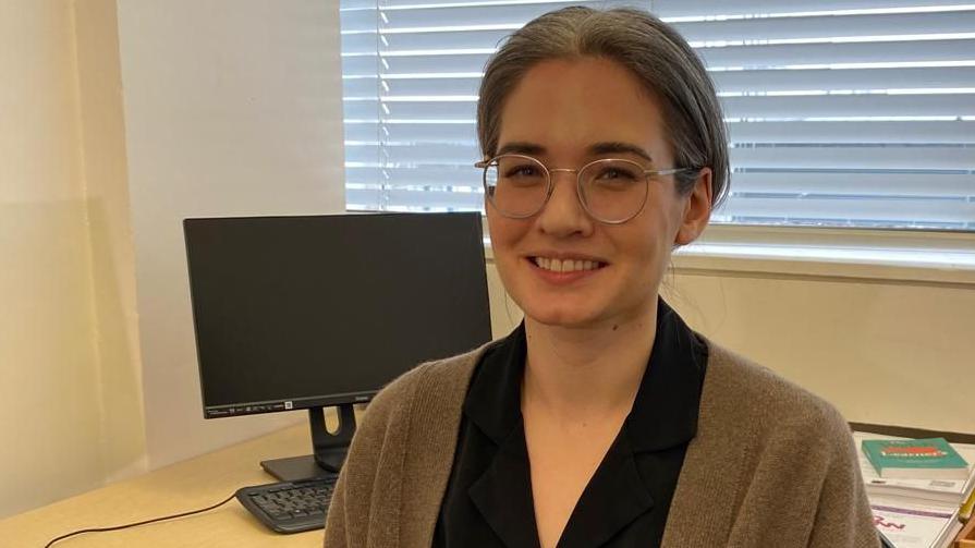 Dr Robyn Jackowich is sitting in her office, in Cardiff University, with her hair tied back - and wearing round silver glasses frames. She is smiling at the camera, sitting at her desk.