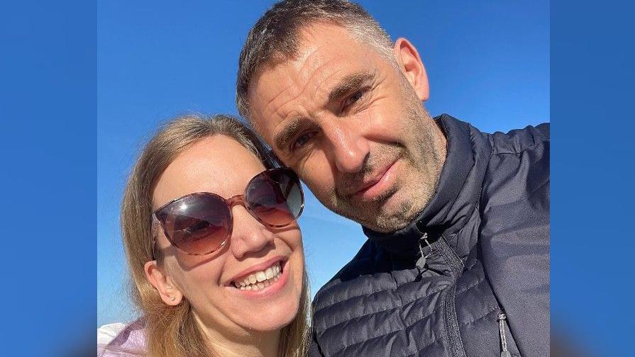 Jess Maggs, on the left, smiling at the camera wearing sunglasses with Rob Maggs on the right. Rob is wearing a black coat.