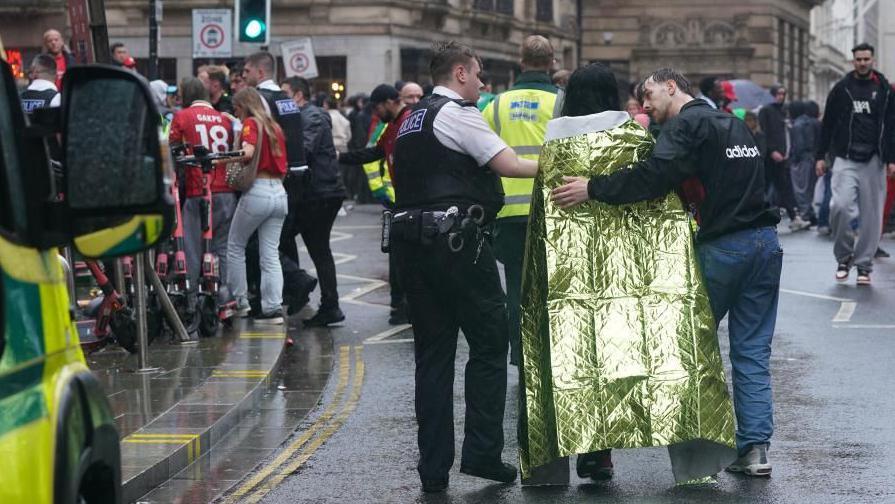 A police officer and man comforting a woman who is draped in a first aid blanket