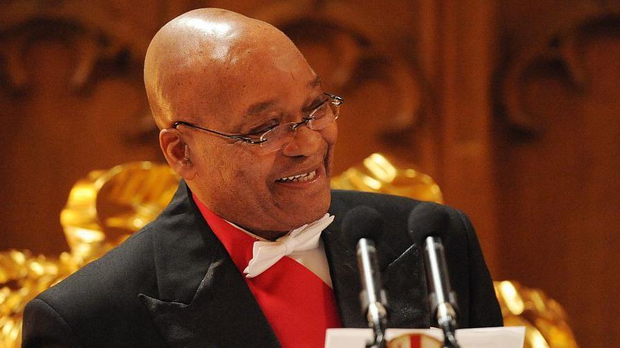 A head and shoulders image of Jacob Zuma in a white bow tie and standing at two microphones. He is looking to his left and is smiling.