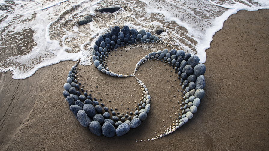 Waves beginning to crash on the beach hitting another art piece made up of three interlocking rock and shell formations that connect in a spiral.