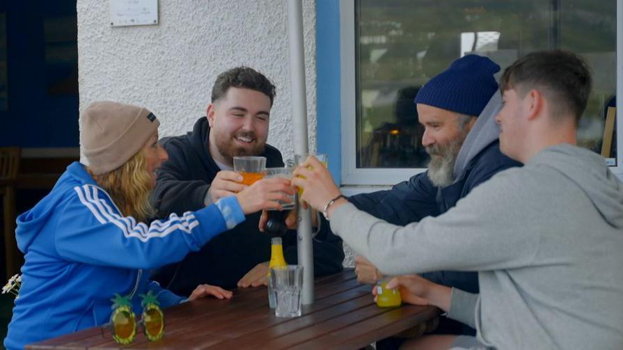 Jules, wearing a blue jacket and beige hat, taps her glass in a cheers with her son Benny next to her, son Chevy across from her and husband Greg. They are sitting outside on a picnic table next to a building.