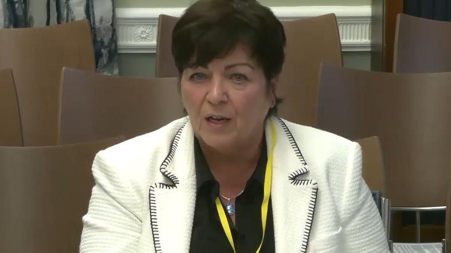 A head shot of Rita Devlin. She has short brown hair. She is wearing a white blazer with a black shirt. Behind her are rows of brown chairs. She has a yellow lanyard around her neck.