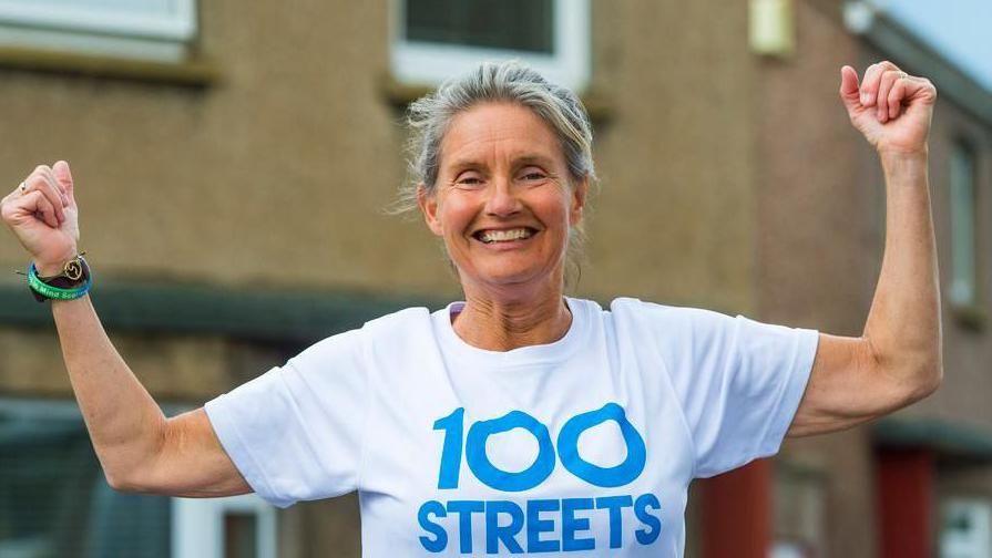 Jenny Hastings is standing in a street of harled brown houses. She is smiling broadly, holding her arms in the air triumphantly. She is wearing a blue on white '100 streets' T-shirt.