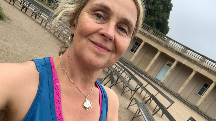 Katie Treutler, wearing a blue running vest, smiles at the camera in a selfie taken in Eaton Park, Norwich, with park buildings and benches behind her.