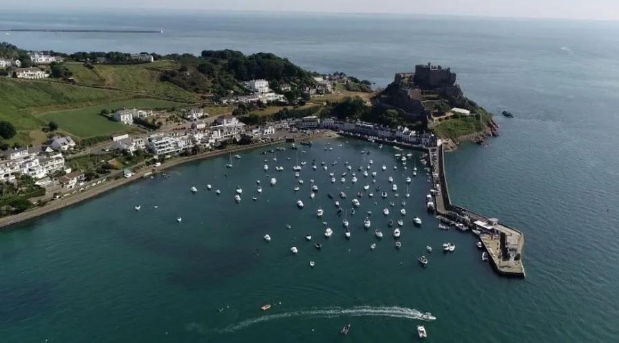 An aerial view of a harbour and a pier with white buildings and green fields in the background.