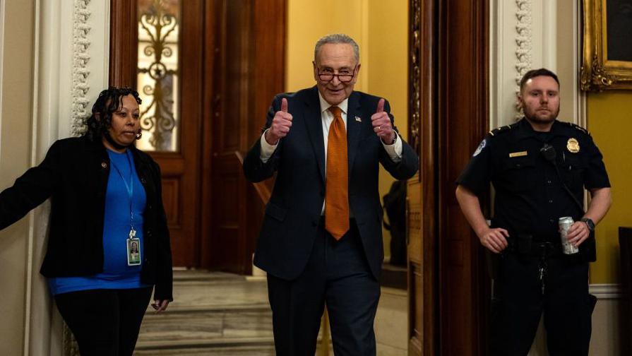 Schumer gives a double thumbs up as he walks out of an ornate doorway opened by a female worker on the left and guarded by a male police officer on the right