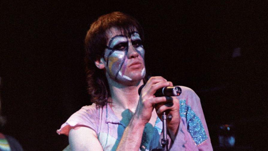 Fish from the band Marillion. He is on stage, holding a microphone. His hair is in a mullet style and his face is covered in white and blue face paint. He is wearing a pink T-shirt with blue glitter on it and is glancing sideways at the camera.